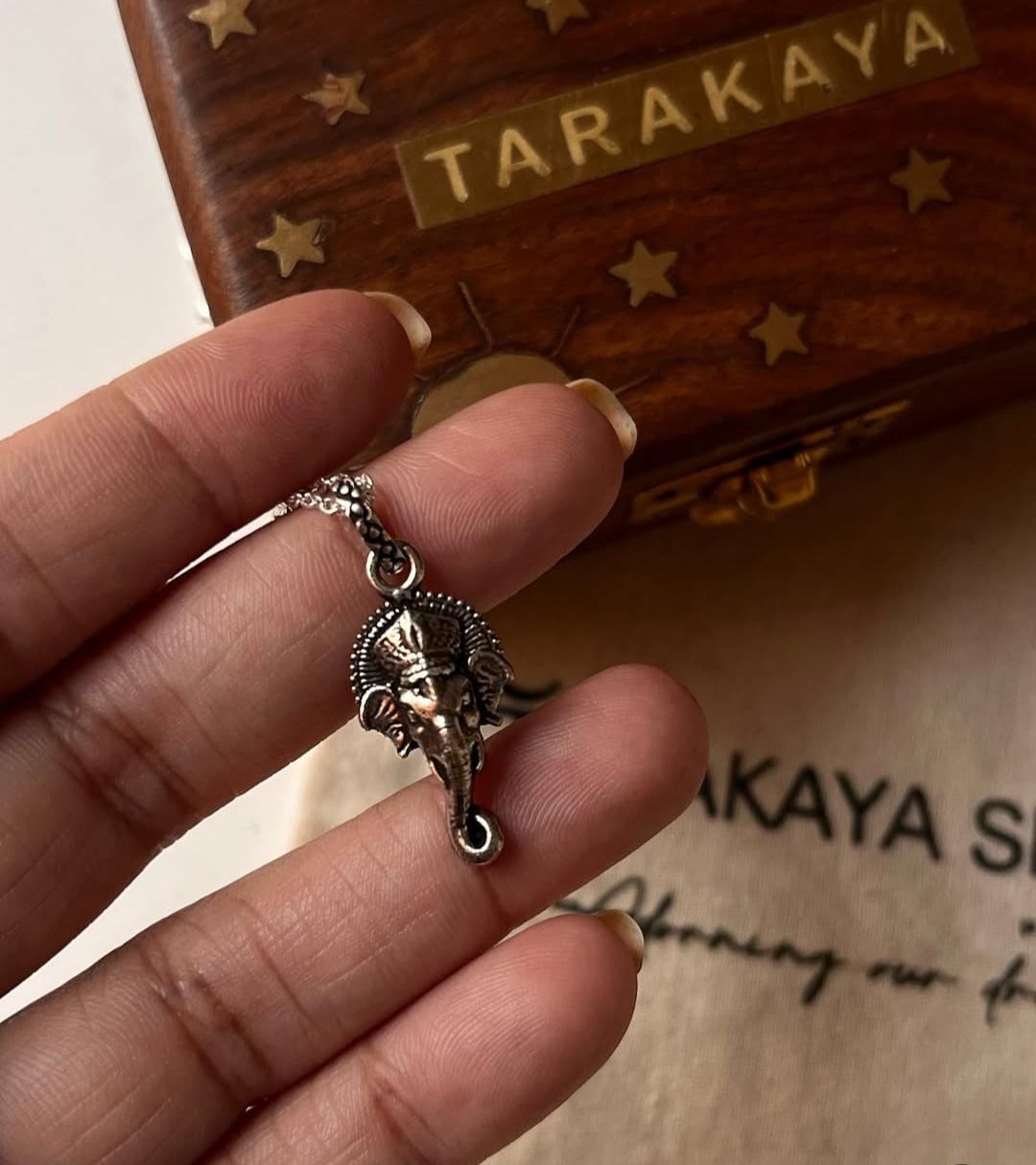 Hand holding a Ganesha face silver pendant, showcasing detailed craftsmanship and design against a decorative background.