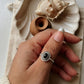 Hand wearing a Black Onyx Rawa Tribal Silver Ring, surrounded by seashell and wooden items on a soft fabric surface.