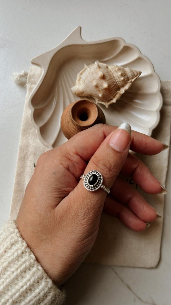 Hand wearing a Black Onyx Rawa Tribal Silver Ring, surrounded by seashell and wooden items on a soft fabric surface.