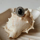 Black Onyx Rawa Tribal Silver Ring on a seashell, showcasing its deep black color and intricate silver detailing.
