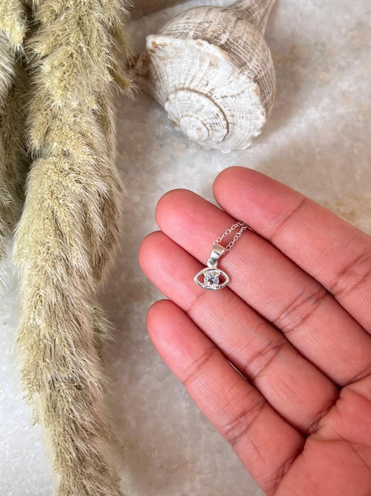 Hand holding a Blue Topaz Tiny Evil Eye 925 Sterling Silver Necklace on a textured surface.